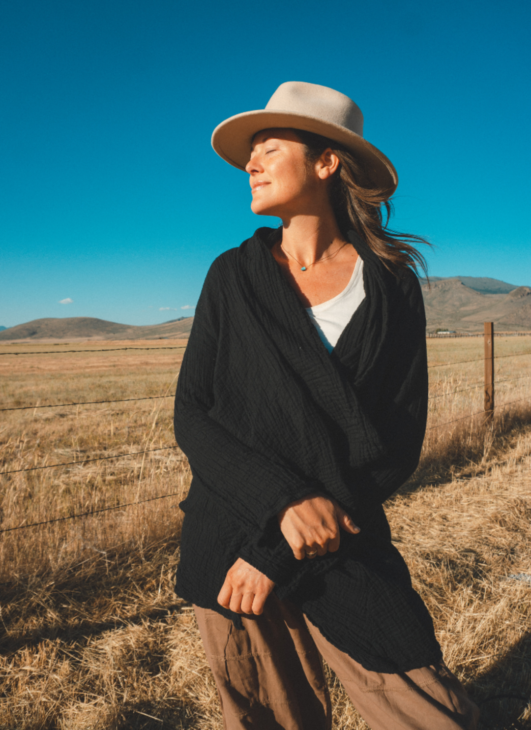 Woman stands in sun wearing tan felt hat, black wrap over white tank top and brown pants.