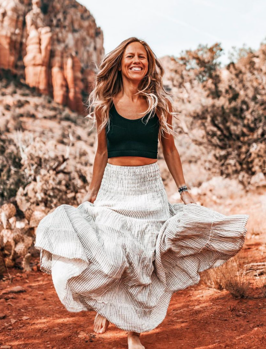 Barefoot female model earring black tank crop top and long white skirt with blue pinstripes.