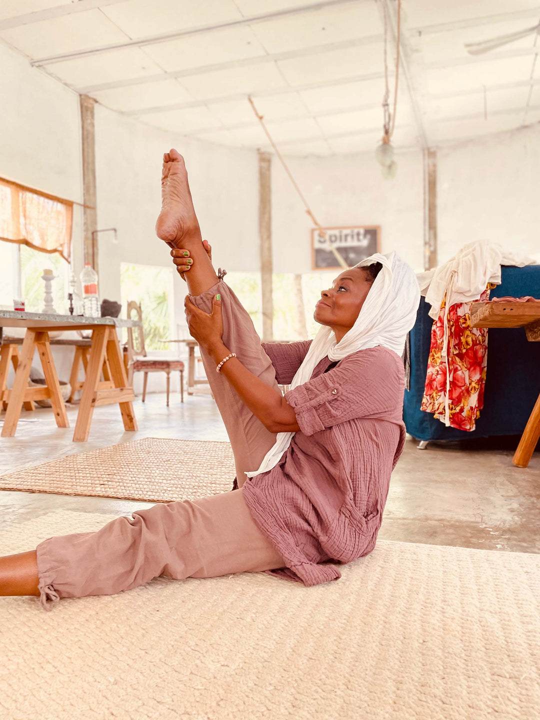 Woman sits on floor stretching while wearing white head scarf, pink tunic top, and brown pants.