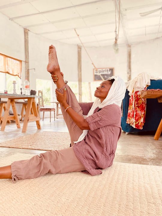 Woman sits on floor stretching while wearing white head scarf, pink tunic top, and brown pants.