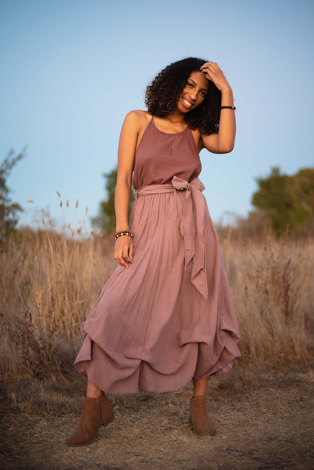 Female model with curly dark hair stands outside. Her left hand is on her head and she is smiling. She wears a rose-color tank top and long tiered skirt with ankle-heigh brown boots.