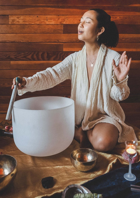 Woman wearing cream robe sits on floor with singing bowls.