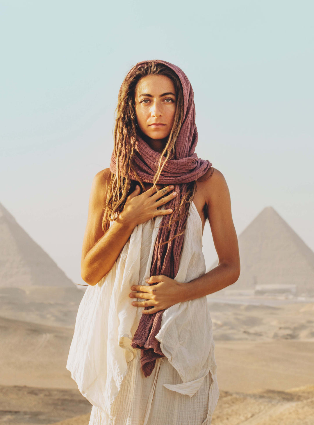 Woman with dreadlocks has head covered with maroon scarf. She is wearing an un-dyed flowing tank top and natural bottoms. Her right hand is on her heart.