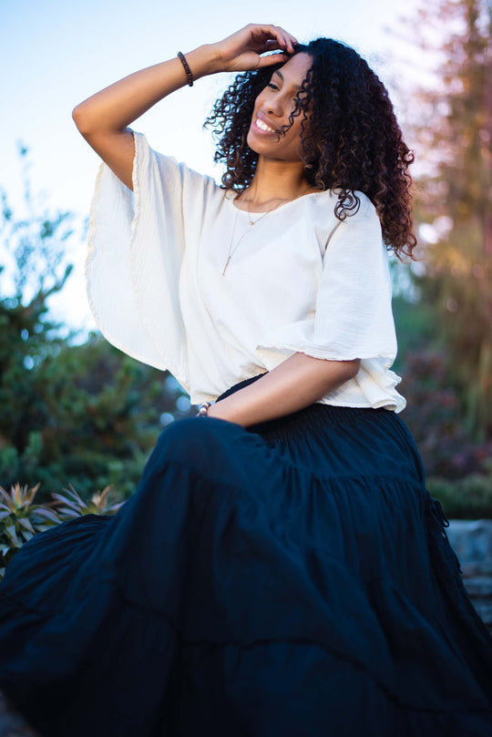 Woman dressed in flowy un-dyed shirt and black maxi skirt sits on stone steps. Her right hand is in her hair.