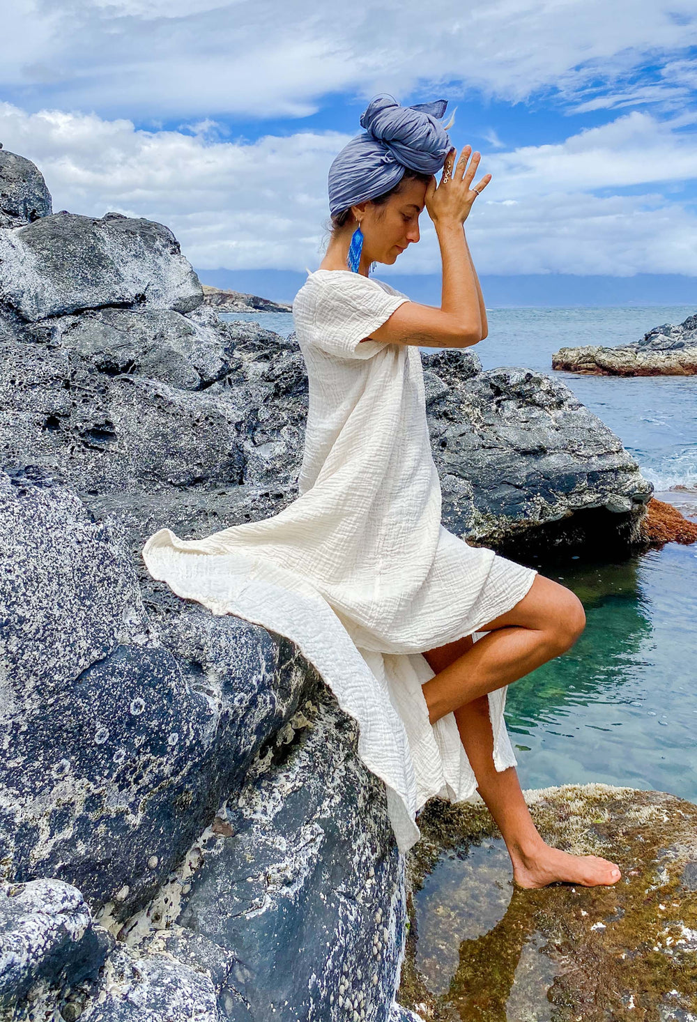 Female model sits a water's edge on a rock wearing blue head scarf and long un-dyed dress.