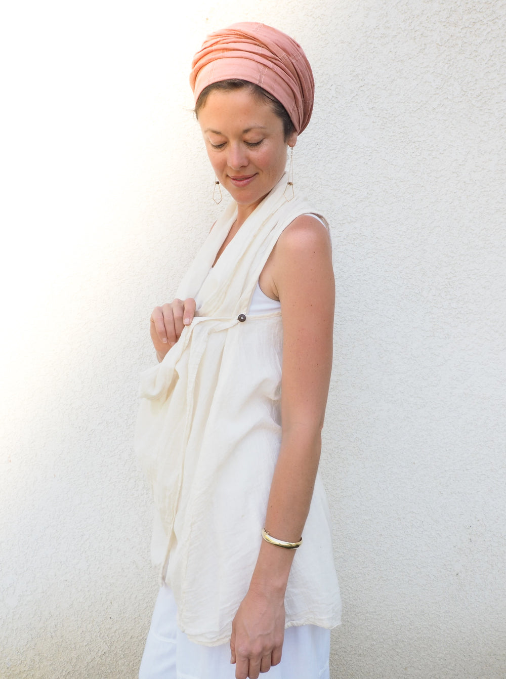 Female model wears peach head wrap, un-dyed vest over a white shirt and white pants. She is showing the button on the best. She stands against a white stucco wall.