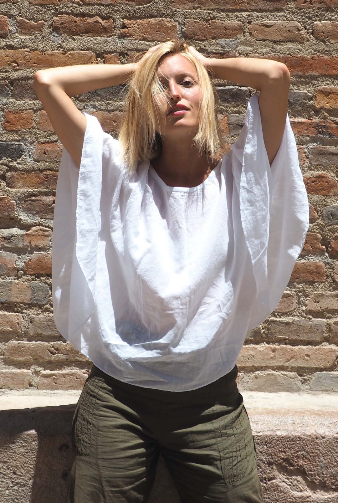 Woman stands in front of brick wall dressed in white shirt with flowing, open sleeves and olive green pants.