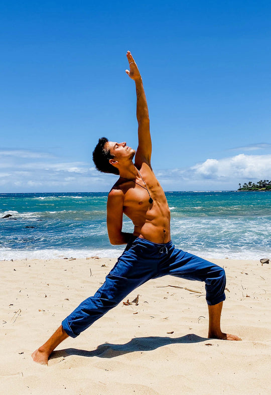 Active male model is shirtless and wearing blue athletic pants. He is in Peaceful Warrior yoga pose. In the background is sand and water.