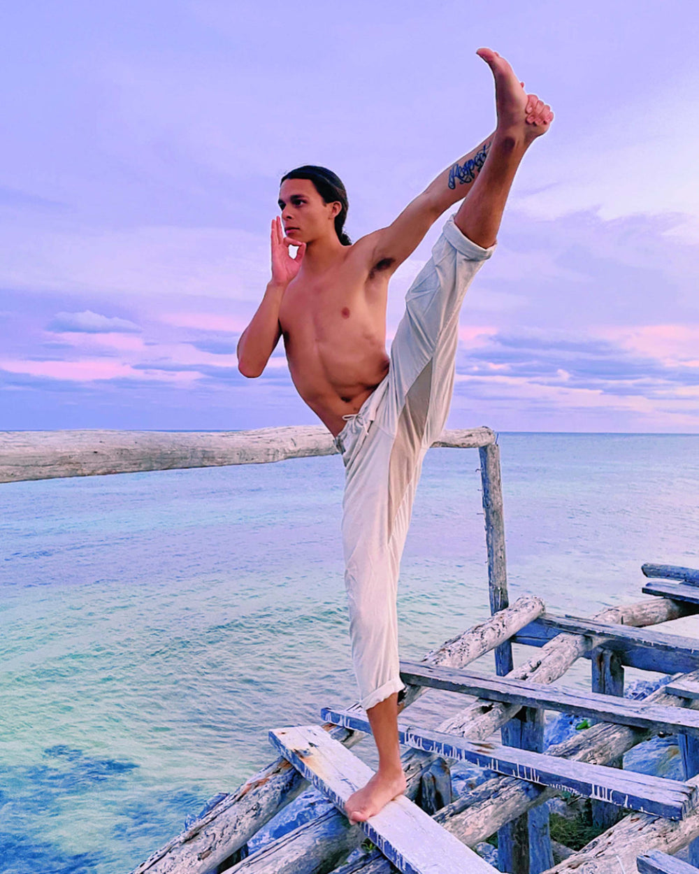 Male model is shirtless and balancing on right leg with left leg extended up above his head. He is holding his left foot with his left hand. His right hand is making a sign near his face. He is wearing natural pants. He is balancing on a board above water.