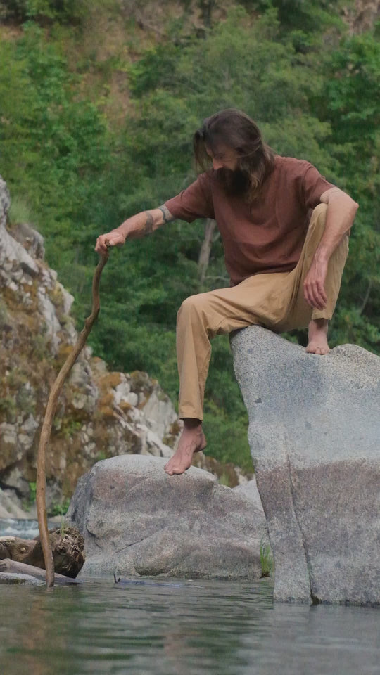 Man is wearing rust-color shirt and tan pants. He sits on a rock in the water, dipping a long stick into  the water.