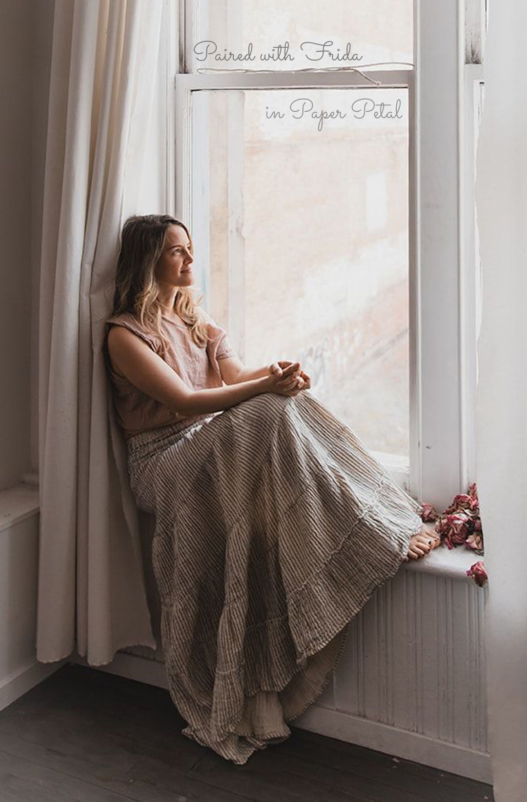 Female model sits on ledge of window sill with bare feet. She is smiling and looking out the window with hands resting on her knees. She is wearing a light pink top and long white skirt with blue pinstripes.