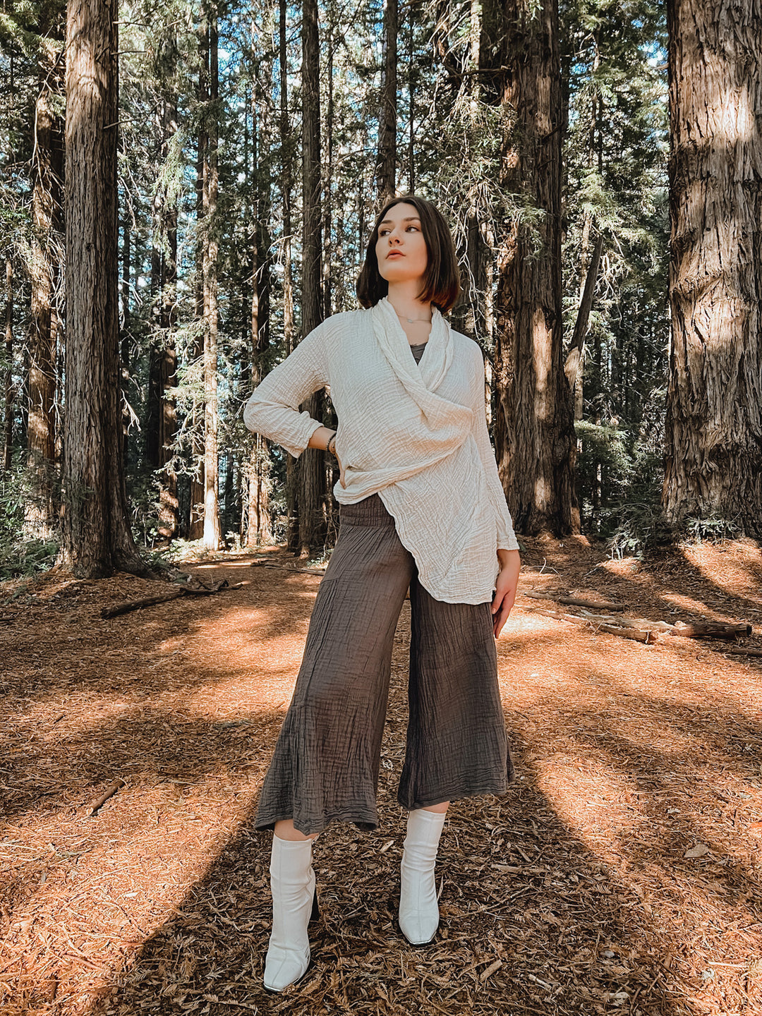 Female clothing model is adorned in cream cloak, brown flowing pants and white boots. She stands in a forest.