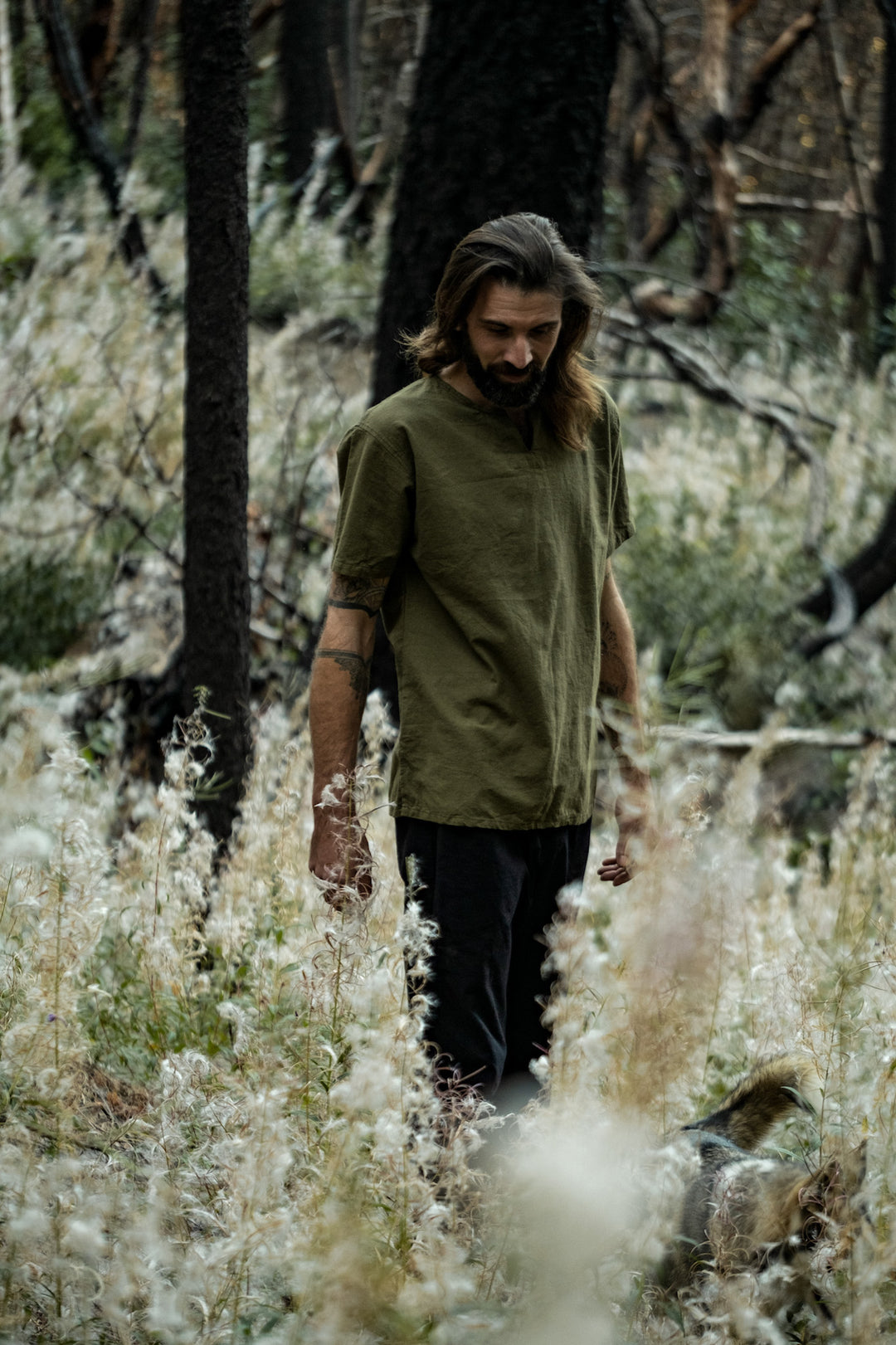 Model in short-sleeve olive green cotton shirt and black bottoms standing in tall flowers.