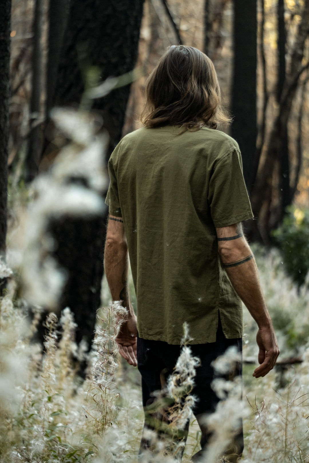 Back side of person in olive green shirt with short sleeves and black pants walking through forest and plants.