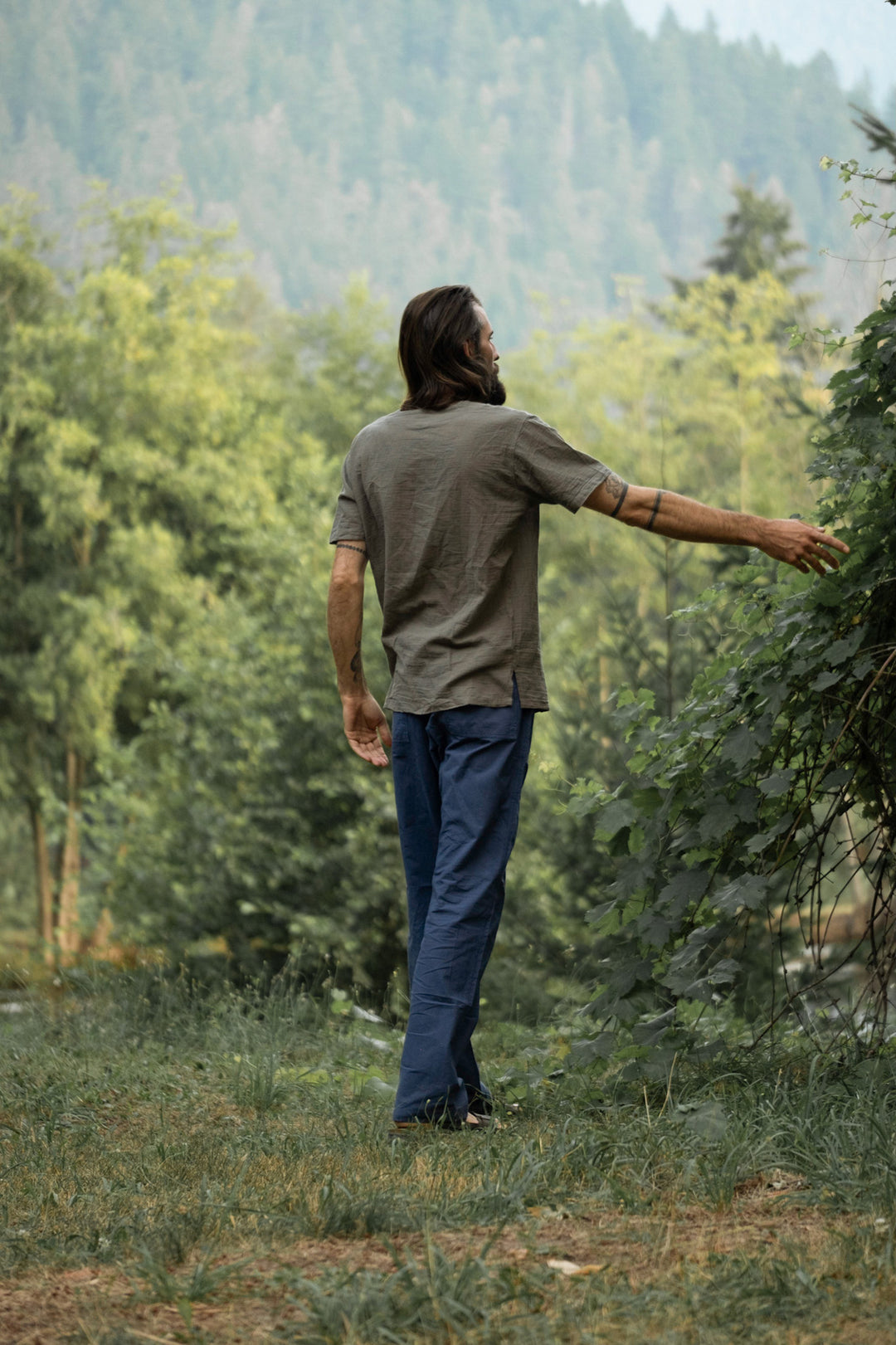 Model walks through forest dressed in short-sleeved shirt and blue pants.