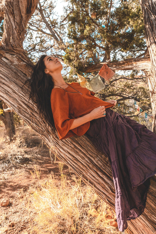 Woman relaxes against tree trunk in a red flow top and purple skirt.