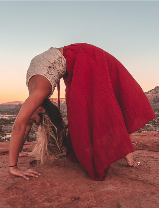 Female model is doing a back bend on red rock with mountain vista in background. She is wearing lacy white tank crop top and long red shirt. She is barefoot.