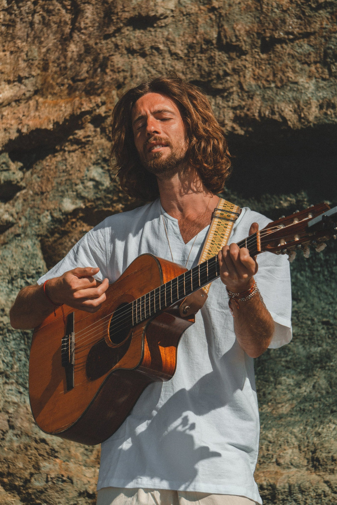 Man with guitar stands against large rock dressed in a white gauze shirt and light-color pants.