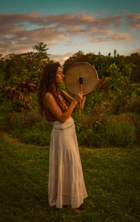 Woman stands in green grass with drum dressed in a natural white maxi skirt.
